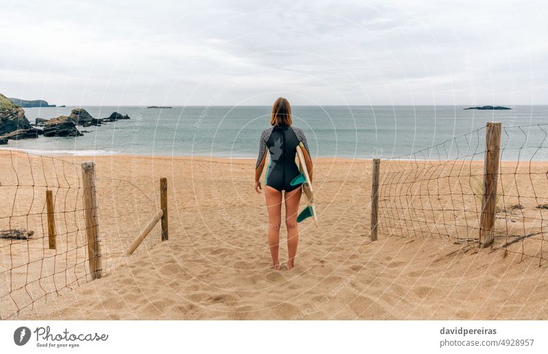 Surfer woman with wetsuit and surfboard looking at the sea unrecognizable young surfer back sand beach swim carrying winter people 20s outdoors sport cold