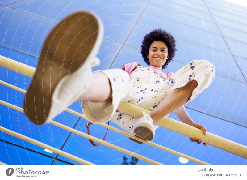 Smiling black woman sitting on railing fence street city urban appearance style apparel positive building glad optimist smile content cheerful garment sneakers