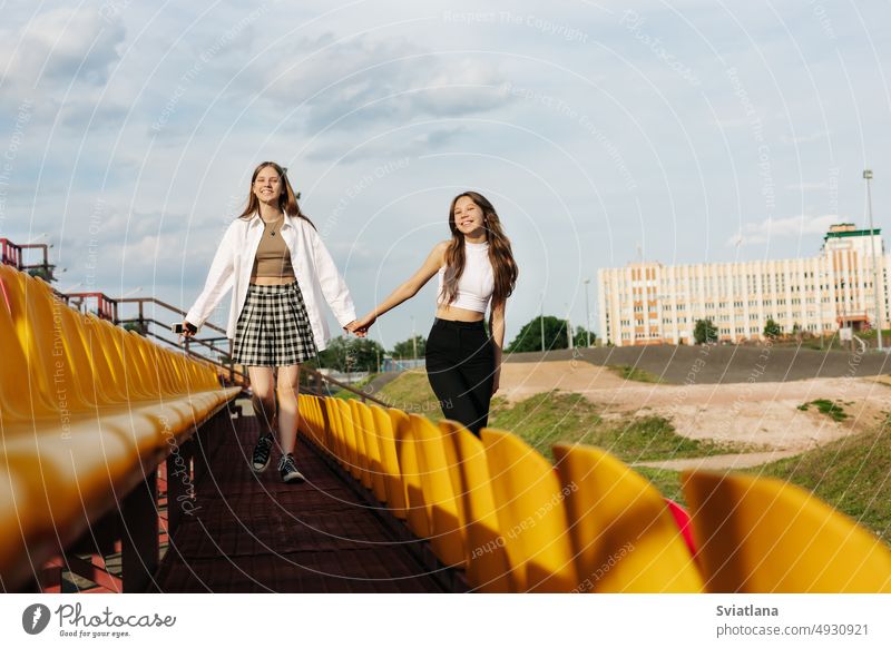 Two teenage girls walk together through the stands of the school stadium, talking, holding hands, best friends return home after training friendship student