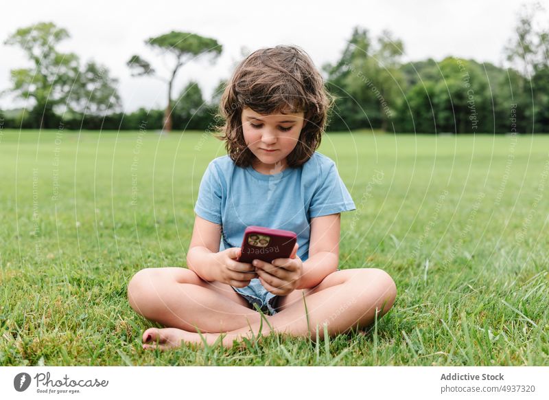 Happy girl using smartphone on park lawn watch video weekend summer pastime happy child gadget device cellphone delight grass glad internet childhood online kid