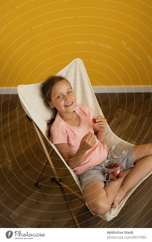 Cheerful girl eating strawberries on deckchair strawberry smile home weekend summer happy fresh kid jar casual cheerful legs crossed sweet healthy food daytime