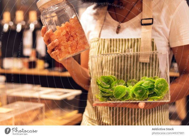 Anonymous female vendor with dried fruits woman show container zero waste store portrait young ethnic uniform box jar pineapple slice kiwi piece shop grocery