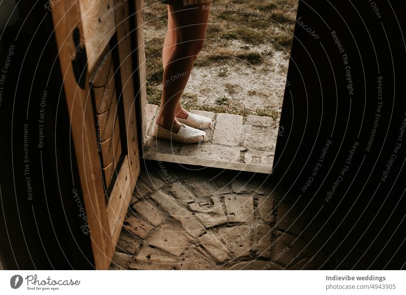 A woman is standing outside the threshold of a door. Rustic vintage stone brick pavement inside and outside. Beautiful hand carved door. Dark copy space.