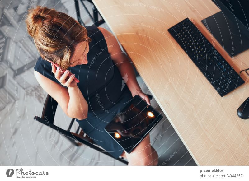 Businesswoman having business call working on tablet in office. Mature busy woman using touch pad computer standing by window in modern interior smartphone talk