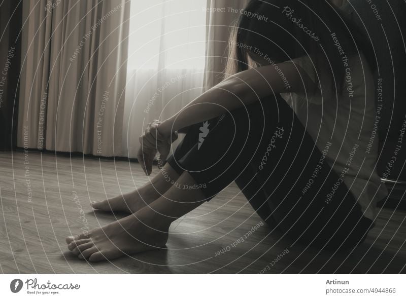 Depressed and stressed woman sitting on the floor near curtains in bedroom. Sad woman sitting in the dark room. Girl with mental health problems. Unhappy life. World Mental Health Day concept.