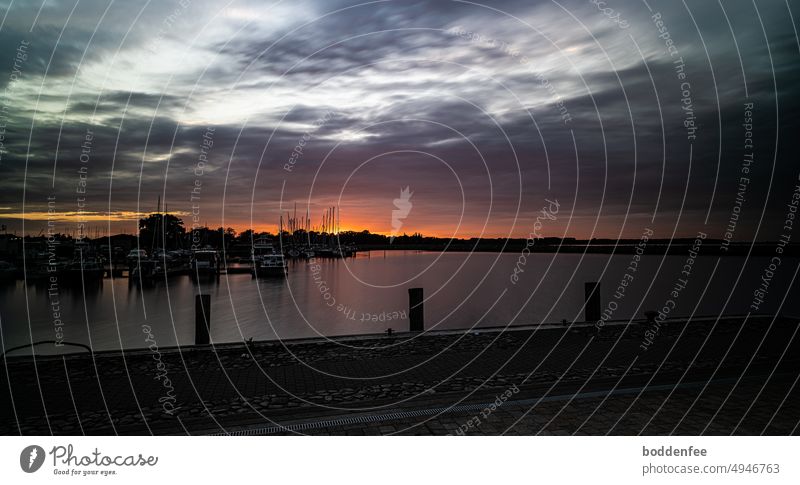 Summer evening at Barth harbor, sunset in August, taken with ND filter, in the foreground a piece of the quay with 3 bollards, in the left center of the picture motor and sail boats, in the background yellow-red sky with black clouds and bright cloud holes