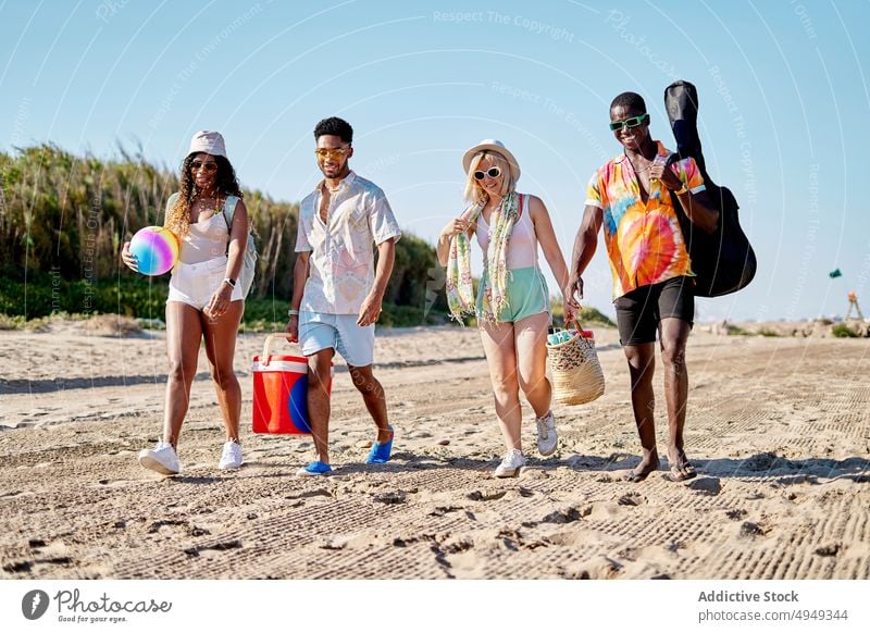 Happy diverse friends walking on beach smile summer weekend together sand happy blue sky resort daytime men glad women young delight stroll group cloudless sky