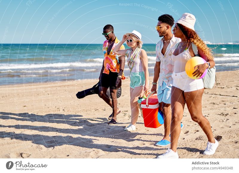 Diverse friends walking on beach summer weekend together sand blue sky resort daytime men women young stroll group cloudless sky bag guitar ball casual diverse