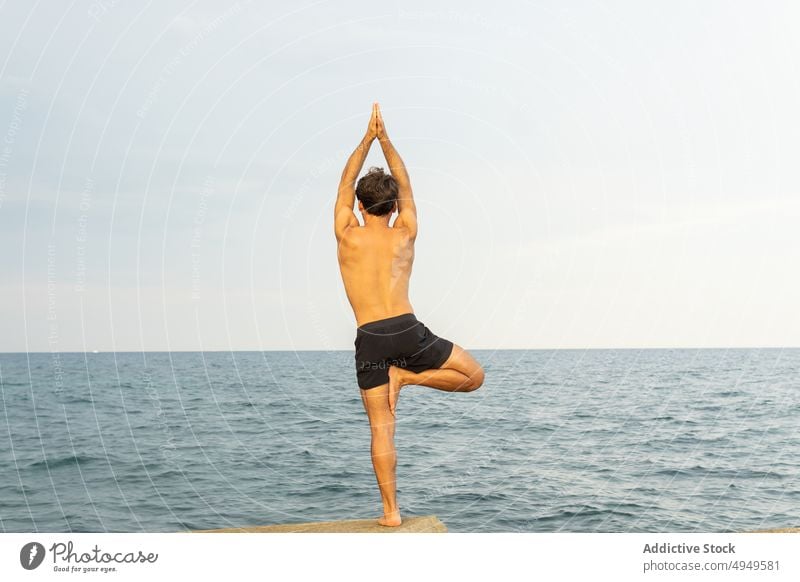 Anonymous man doing Tree pose near sea yoga embankment session tree pose balance rock water practice male stress relief sky gray zen shore asana wellbeing