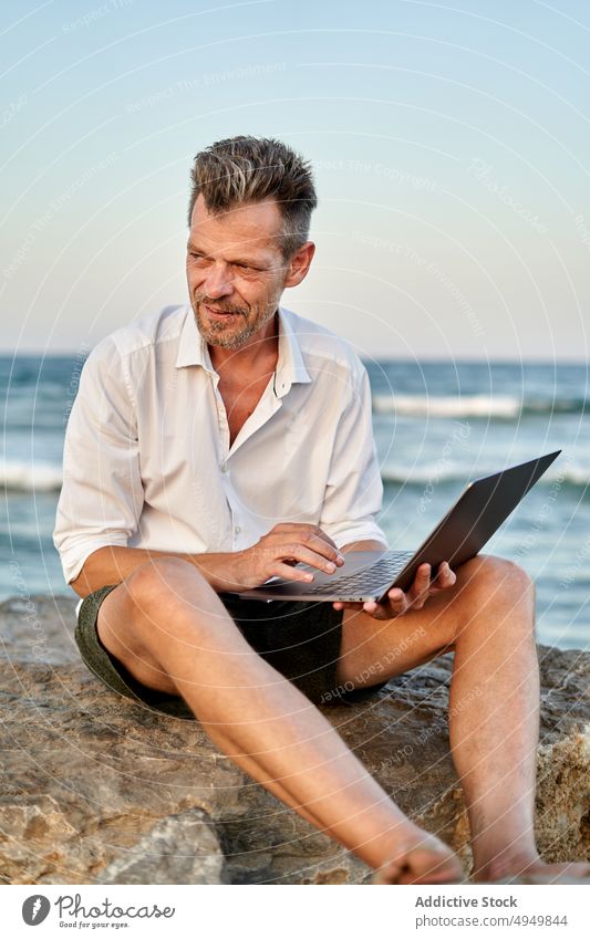 Mature businessman working on rocks near sea laptop using smile data telework male mature middle age independent project sunset happy smart casual positive