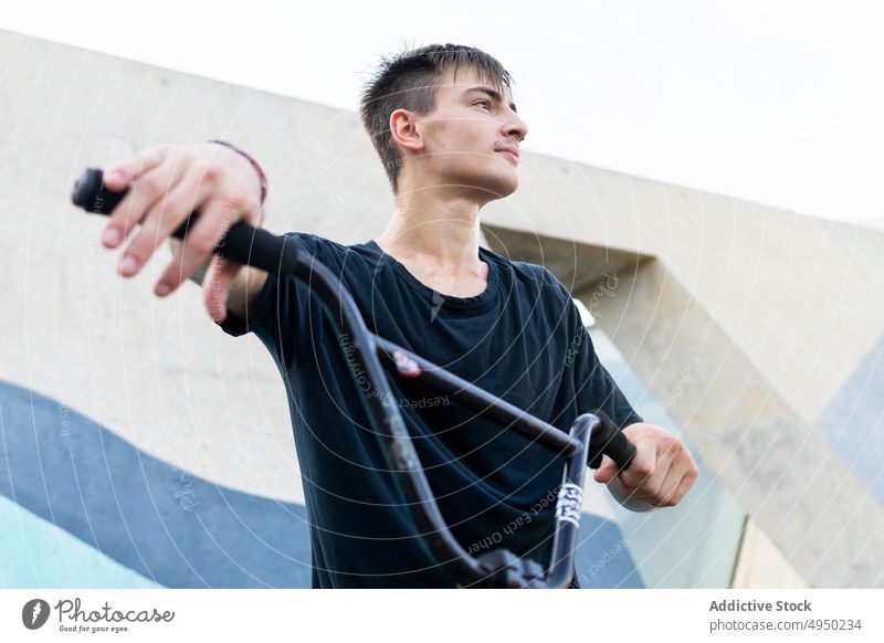 Young man with bicycle in skate park wall hobby weekend break daytime modern urban male young casual t shirt summer bike handlebar touch pause building cyclist