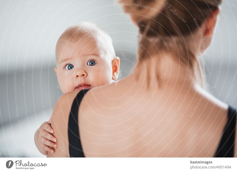 Portrait of sweet baby resting in mothers arms, looking at camera, touching mama shoulder. New mom holding little kid, embracing child with tenderness, love, care. Motherhood concept.