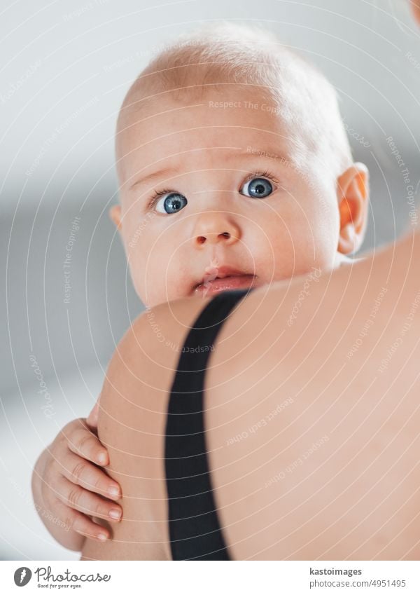 Portrait of sweet baby resting in mothers arms, looking at camera, touching mama shoulder. New mom holding little kid, embracing child with tenderness, love, care. Motherhood concept.