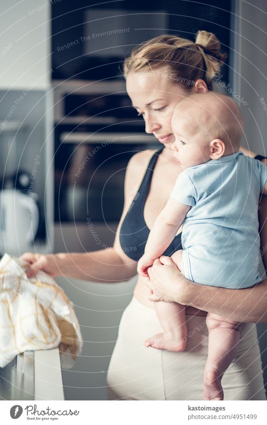 Woman wiping kitchen sink with a cloth after finishing washing the dishes while holding four months old baby boy in her hands. childhood family motherhood woman