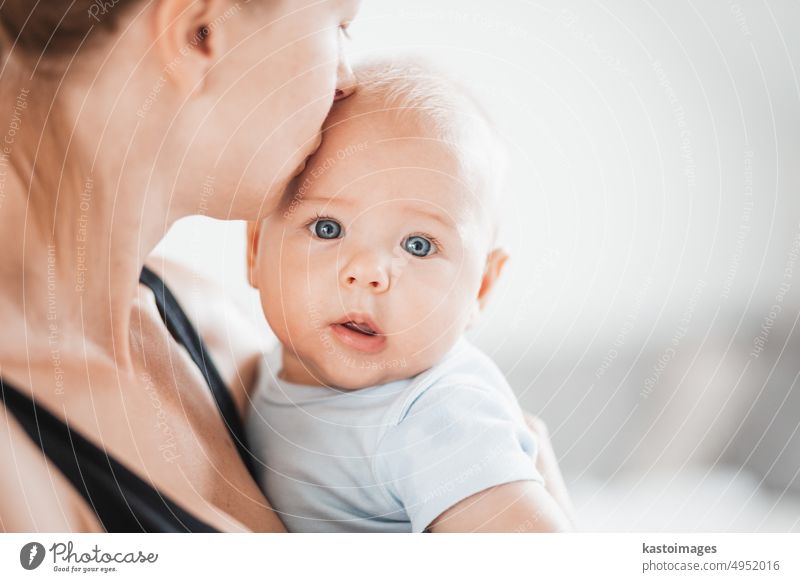 Portrait of sweet baby resting in mothers arms, looking at camera. New mom holding and kissing little kid, embracing child with tenderness, love, care. Motherhood concept.