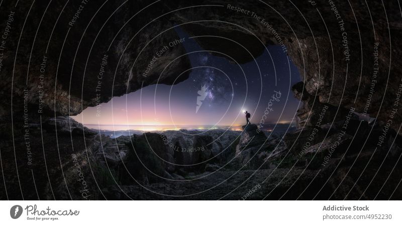 Unrecognizable traveler near cave at night sky starry silhouette entrance stone flashlight dark nature landscape evening countryside tranquil illuminate