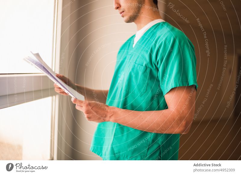 Crop doctor in uniform with paper in clinic hallway medic door profession surgeon protect sterile man window passage hospital professional job corridor light