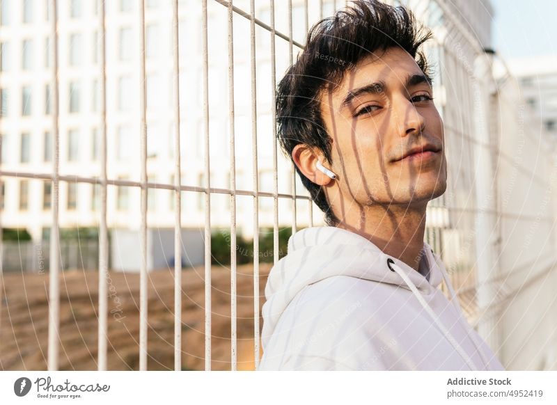 Stylish man standing near metal grid with wireless earphones in city style fence trendy outfit urban content male young street handsome modern lean town delight