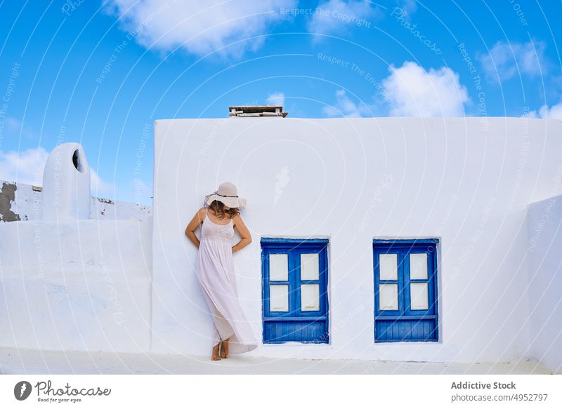 Anonymous woman covering face with hat against rural cottage cover face hide incognito house facade shutter exterior white blue architecture residential