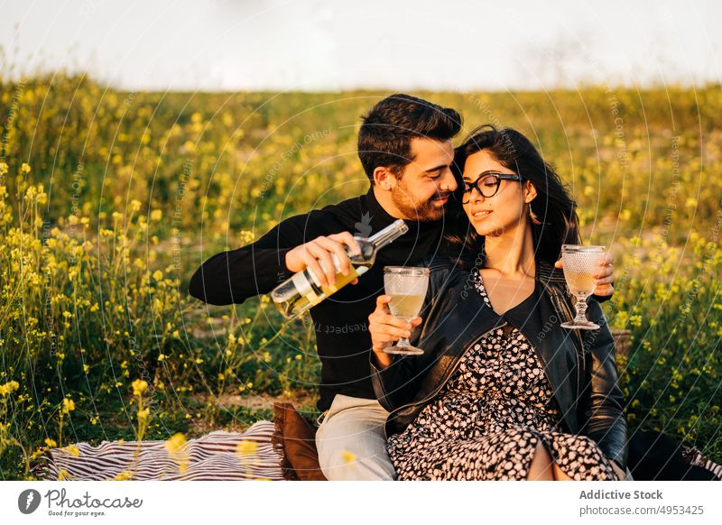 Ethnic couple with bottle of wine on meadow relationship love pour spend time romantic weekend drink beverage alcohol glass bloom flower field sit rest soulmate