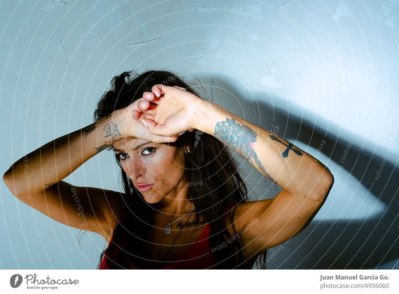 Young tattooed Latina girl with a hard expression and hands crossed on her forehead protects herself from intense light. young latina crossed hands hard shadow