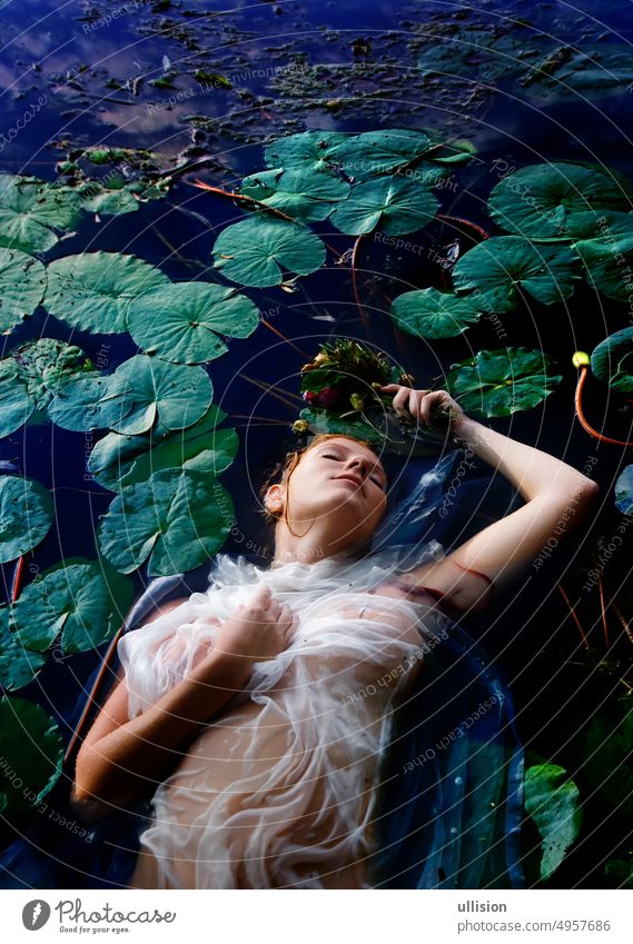 Young sexy unhappy crazy woman Ophelia with curly red hair lies with eyes closed in white dress in the water between water lilies, in the lake, copy space green