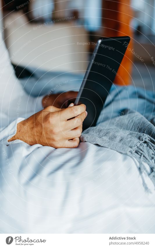 Anonymous man browsing tablet while lying on bed in room using device home online social media surfing connection contemporary male hands young modern comfort