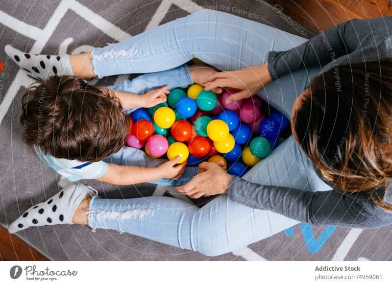 Anonymous mother and son playing with balls on floor sit heap toy colorful nursery home hopscotch mat little woman boy child kid toddler together love game