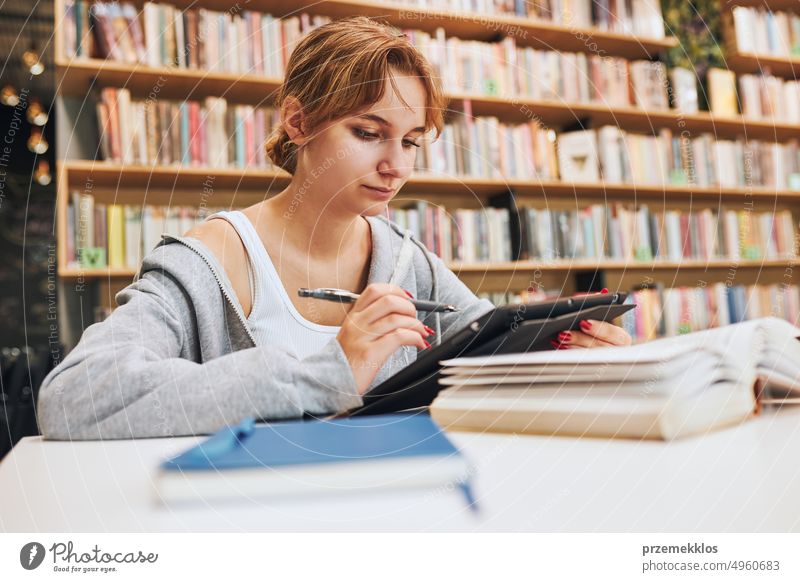 Student learning in university library. Young woman writing essay and making notes using computer tablet. Focused student studying for college exams education