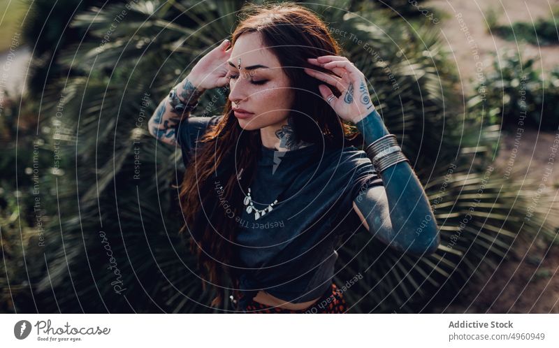 Calm trendy young woman against green tropical plants on flowerbed in park tattoo meditate calm relax street tranquil sensual dream exotic female personality