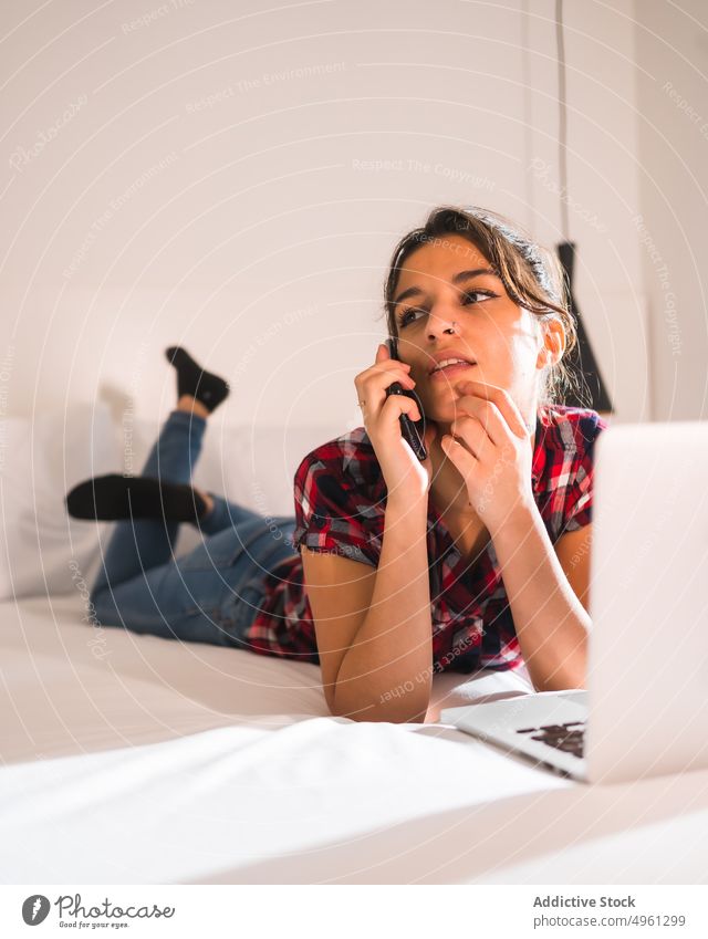 Thoughtful woman speaking on smartphone on bed with laptop conversation touch chin pensive spare time legs crossed using gadget device multimedia cellphone talk