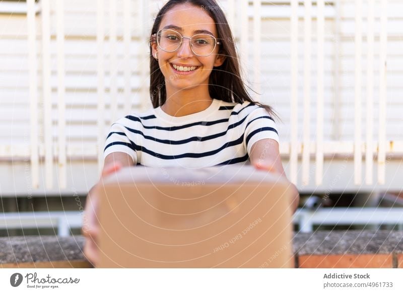 Happy female courier giving carton box and smiling at camera woman delivery give smile shipment order parcel positive happy cardboard young long hair dark hair