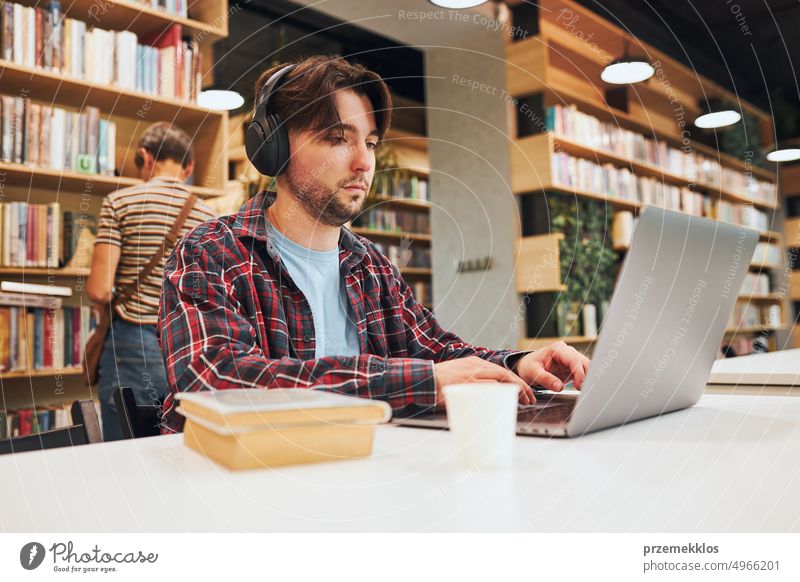 Student learning in university library. Young man preparing for test on laptop. Man listening to online course. Focused student studying for college exams