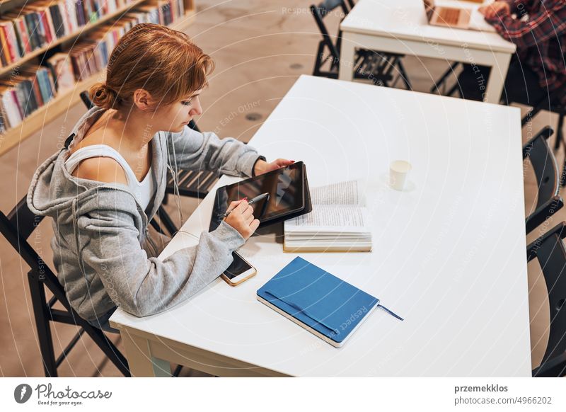 Student learning in university library. Young woman writing essay and making notes using computer tablet. Focused student studying for college exams education