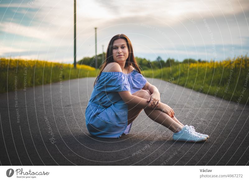 Candid portrait of a shy young girl in a beautiful blue summer dress with white sneakers sitting on the road with a natural breathtaking smile at sunset. Fashion style. European model