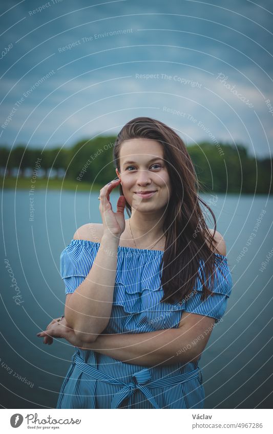 Cute brunette model with long straight dark hair in a summer blue dress with a beautiful realistic smile poses by a lake in the Czech Republic. European woman. Candid portrait with realistic smile