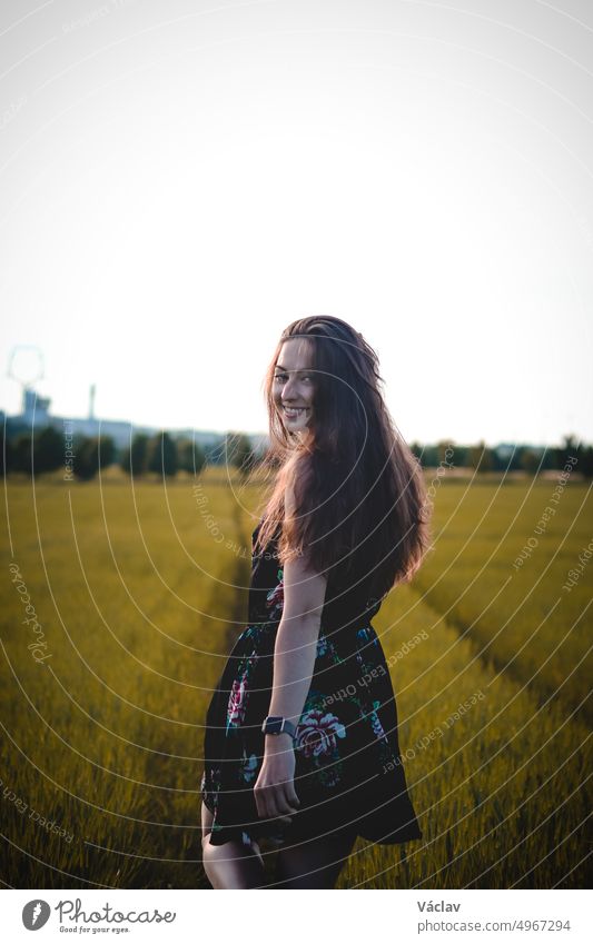 Breathtaking brunette model with a natural wide smile in a dark summer floral dress walks through the tall grass, her wavy dark hair flying. Candid portrait real adult woman. Czech republic, Europe