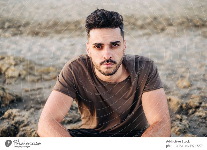 Handsome young Hispanic man sitting on sandy beach clasping hand thoughtful contemplative brunet resting folding relaxing ethnic hispanic waiting thinking