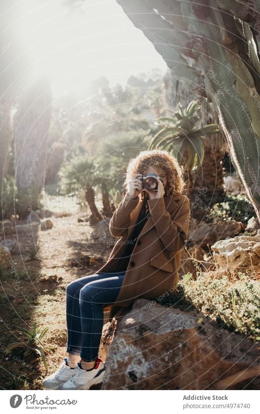 Portrait of beautiful curly blonde woman using a retro camera while sitting on rock portrait nature outdoors accessory apparel clothing coat confident cute