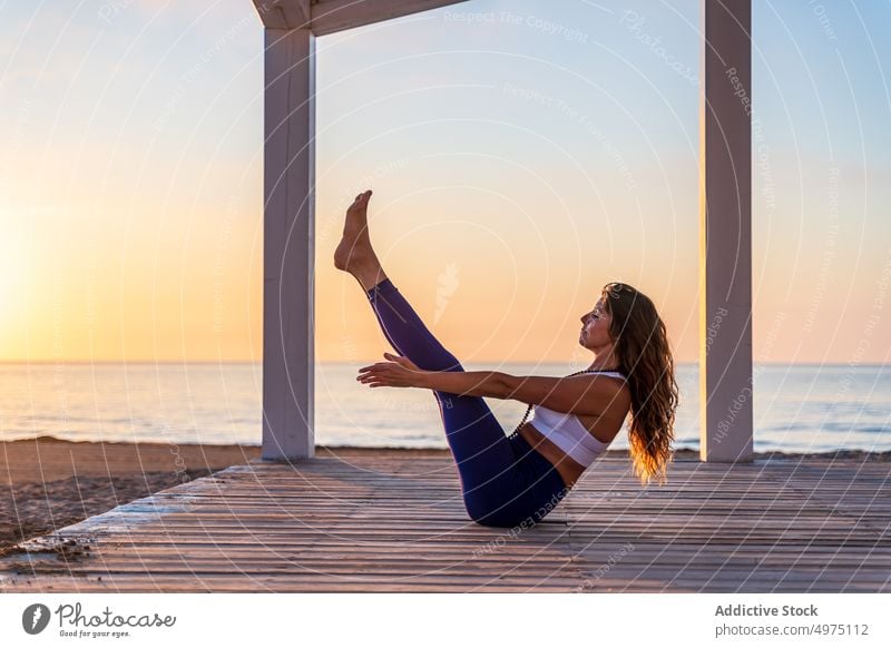 Focused woman practicing yoga in Boat pose on seashore seaside sunrise practice boat navasana balance focus activewear slim healthy wooden promenade picturesque