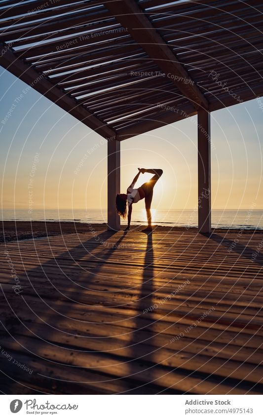 Woman doing yoga in standing split pose at seaside woman balance flexible sportswear seashore sunrise practice healthy harmony wellness slim stretch posture