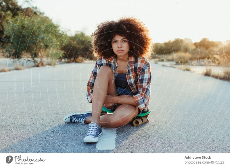 African American woman sitting on penny board on the asphalt road summer skater street cool millennial afro trendy tranquil hipster sunny female ethnic