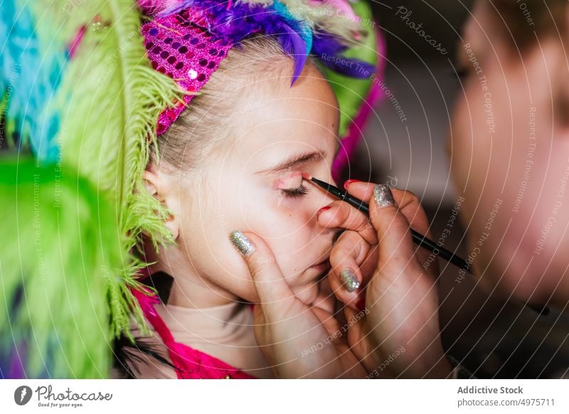 Crop mother applying festival makeup on daughter face carnival prepare airliner home colorful feather headgear girl woman kid child fashion event glamour