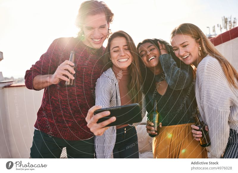 Joyful diverse friends taking selfie during party on terrace smartphone grimace make face beer joyful booze together sofa device v sign two fingers laugh gadget