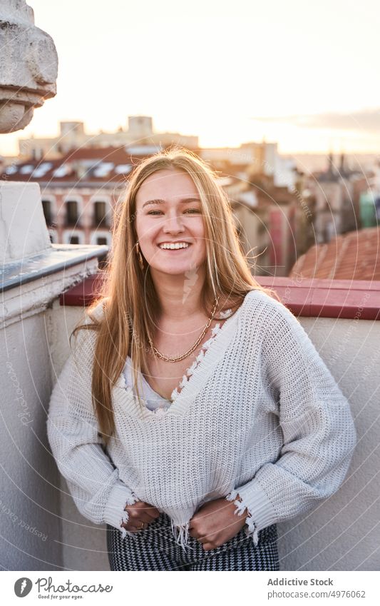 Smiling young woman standing on terrace cheerful toothy smile mood happy rooftop twilight town expressive excited casual content railing building optimist city