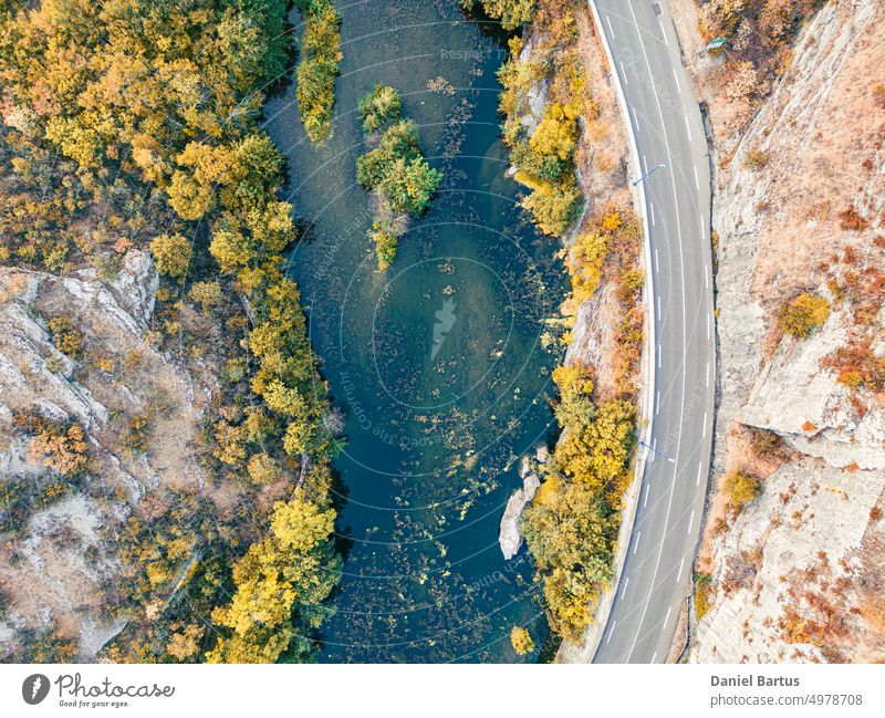 A road on a mountain slope overlooking a river with colorful vegetation and trees on the edge of the slope. Drone flight view. Background autumn background