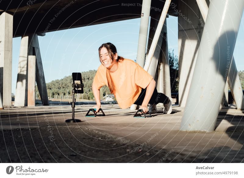 Young woman influencer blogger recording a live streaming on social media with mobile phone,working up with push ups bar cross.Teaching,explaining and doing a demonstration.Training cheap from home.