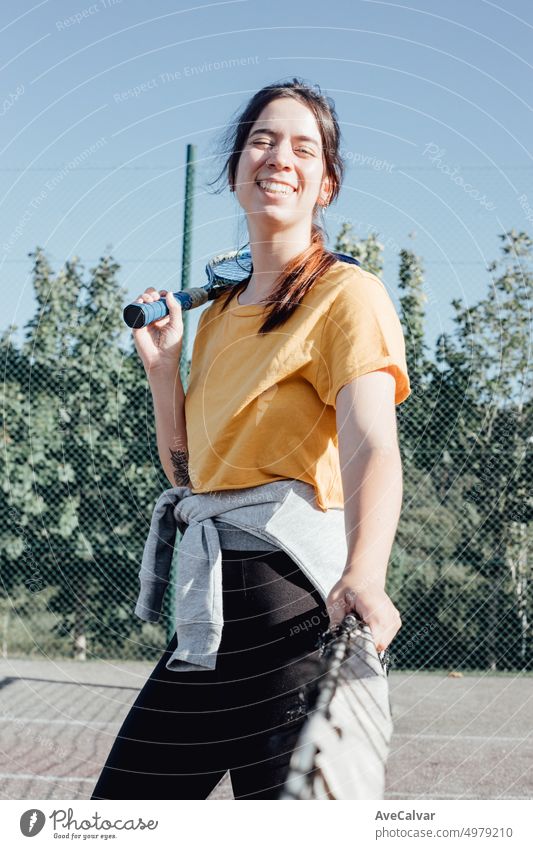 Young girl beginner tennis player waiting leaning on the tennis net to start the match with big smile.Holding a racket excited for learn a sport.New hobby and healthy life and style,happy and nervous