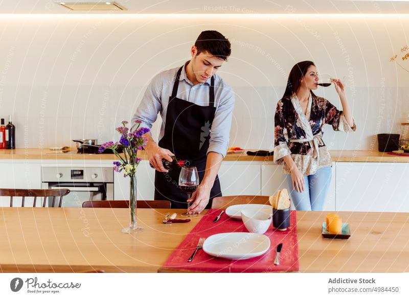 Man pouring wine in glass near woman drinking from glass in kitchen couple pinafore beverage plate bottle table dinning young dinner feast food dish preparation