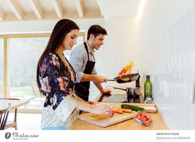 Woman cutting vegetables near man pouring oil on frying pan in kitchen couple preparation hispanic pinafore chopping board bottle carrot woman young attractive